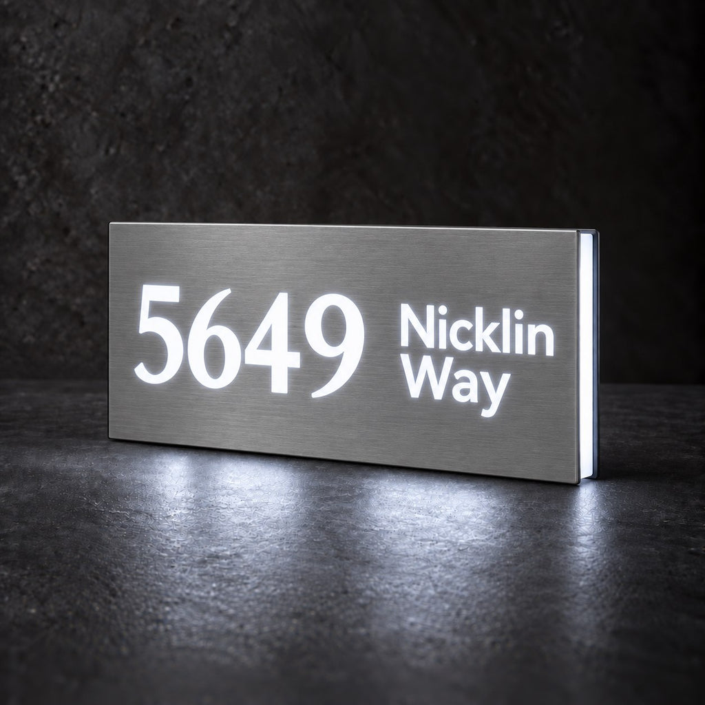 Brushed silver illuminated house number and street name sign with a brushed metal look, The Marcoola, 5649 Nicklin Way cut through and lit from behind in cool white.