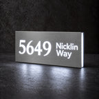 Brushed silver illuminated house number and street name sign with a brushed metal look, The Marcoola, 5649 Nicklin Way cut through and lit from behind in cool white.