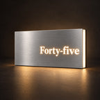 Brushed silver illuminated house number sign with a brushed metal look, The Montville, with the number spelled out as Forty-five, lit from behind in warm white.