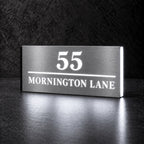 Brushed silver illuminated house number and street name sign with a brushed metal look, The Noosa, 55 Mornington Lane with the number, rule and street name lit from behind in cool white.