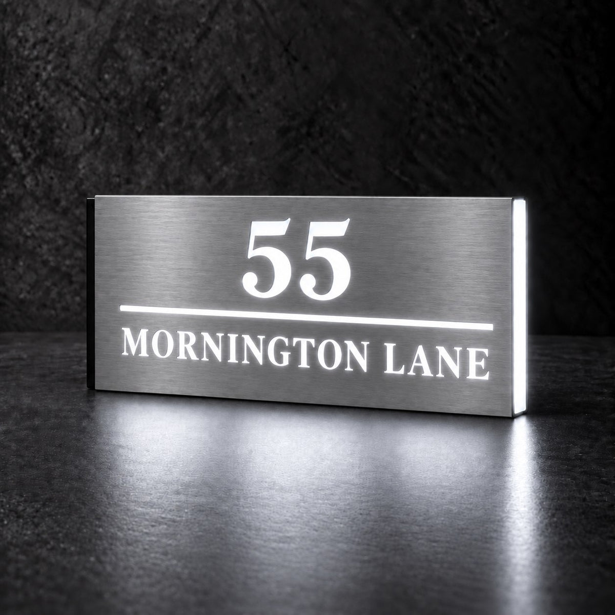 Brushed silver illuminated house number and street name sign with a brushed metal look, The Noosa, 55 Mornington Lane with the number, rule and street name lit from behind in cool white.