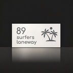 Matte white house number and street name sign, The Yaroomba, reading 89 Surfers Laneway in charcoal lettering, with palm trees and a sun.