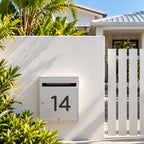 Matte black acrylic house number 14 in the rounded font, fixed flat with double sided tape to the door of a silver letterbox in a white rendered fence.
