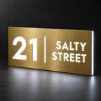 Brushed gold illuminated house number and street name sign with a brushed metal look, The Coolum, 21 Salty Street with the number, rule and street name lit from behind in cool white.