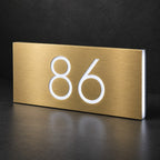 Brushed gold illuminated house number sign with a brushed metal look, The Kawana, displaying 86 in a glowing white LED cutout against a dark background.