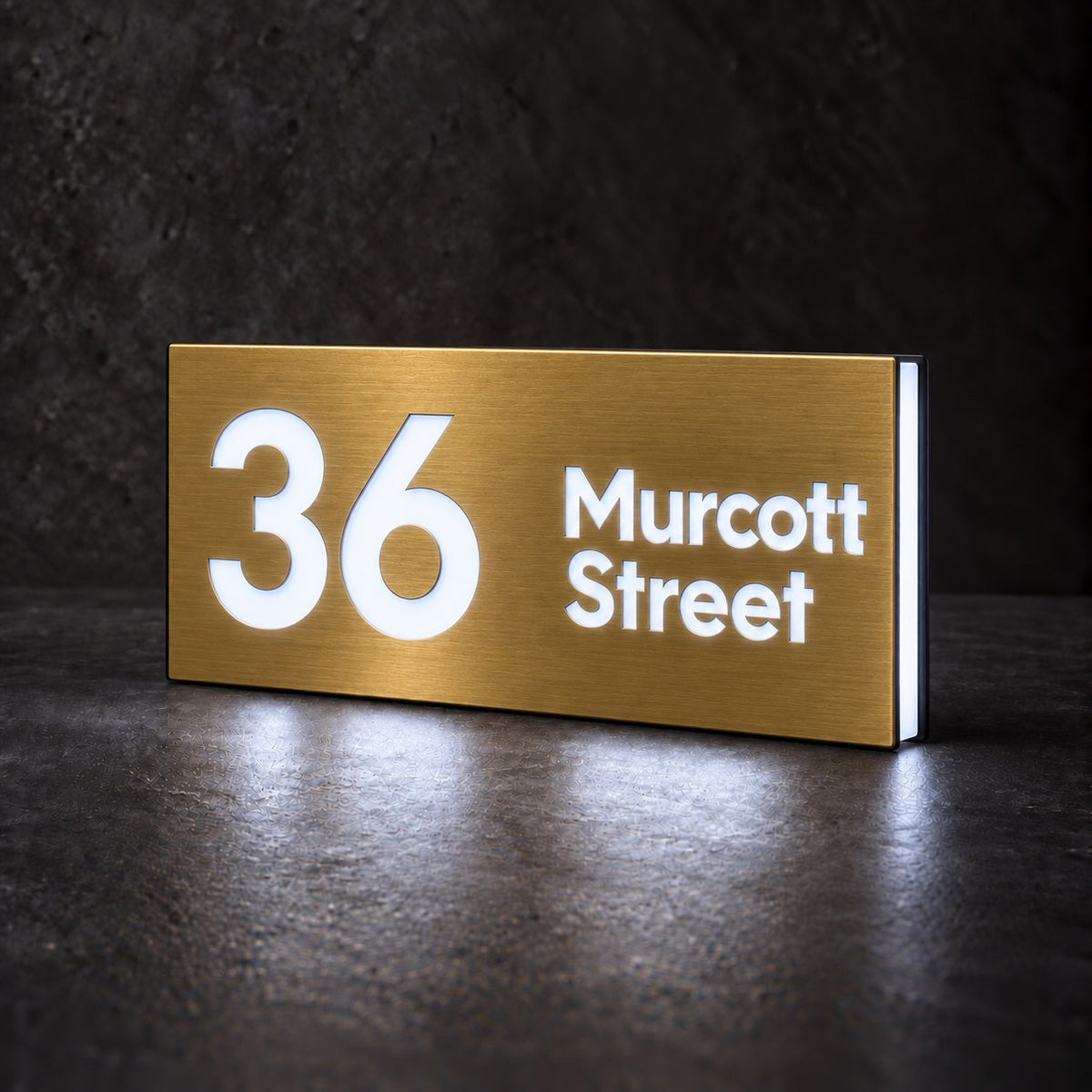 Brushed gold illuminated house number and street name sign with a brushed metal look, The Marcoola, 36 Murcott Street cut through and lit from behind in cool white.