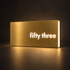 Brushed gold illuminated house number sign with a brushed metal look, The Montville, with the number spelled out as fifty three, lit from behind in warm white.