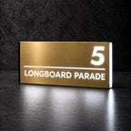 Brushed gold illuminated house number and street name sign with a brushed metal look, The Noosa, 5 Longboard Parade with the number, rule and street name lit from behind in cool white.