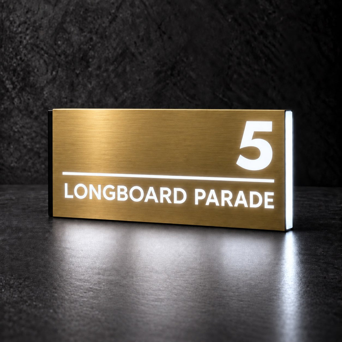 Brushed gold illuminated house number and street name sign with a brushed metal look, The Noosa, 5 Longboard Parade with the number, rule and street name lit from behind in cool white.