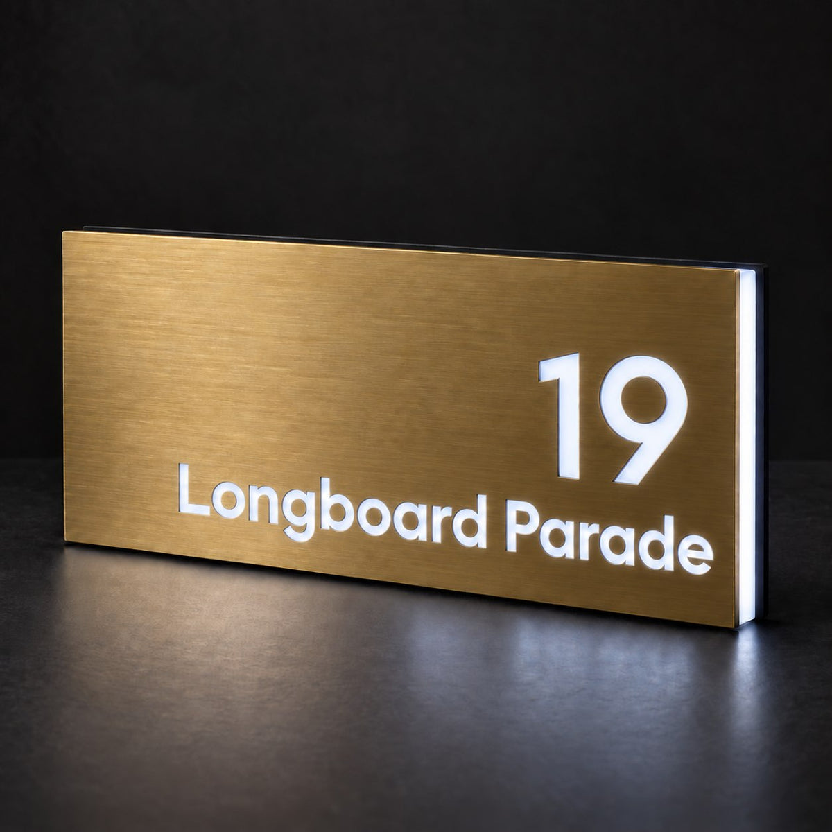 Brushed gold illuminated house number and street name sign with a brushed metal look, The Peregian, 19 Longboard Parade cut through and lit from behind in cool white.