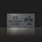 Matte charcoal house number and street name sign, The Yaroomba, reading 65 Bateman Close in black lettering, with palm trees and a sun.