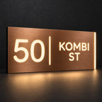 Brushed copper illuminated house number and street name sign with a brushed metal look, The Coolum, 50 Kombi St with the number, rule and street name lit from behind in warm white.