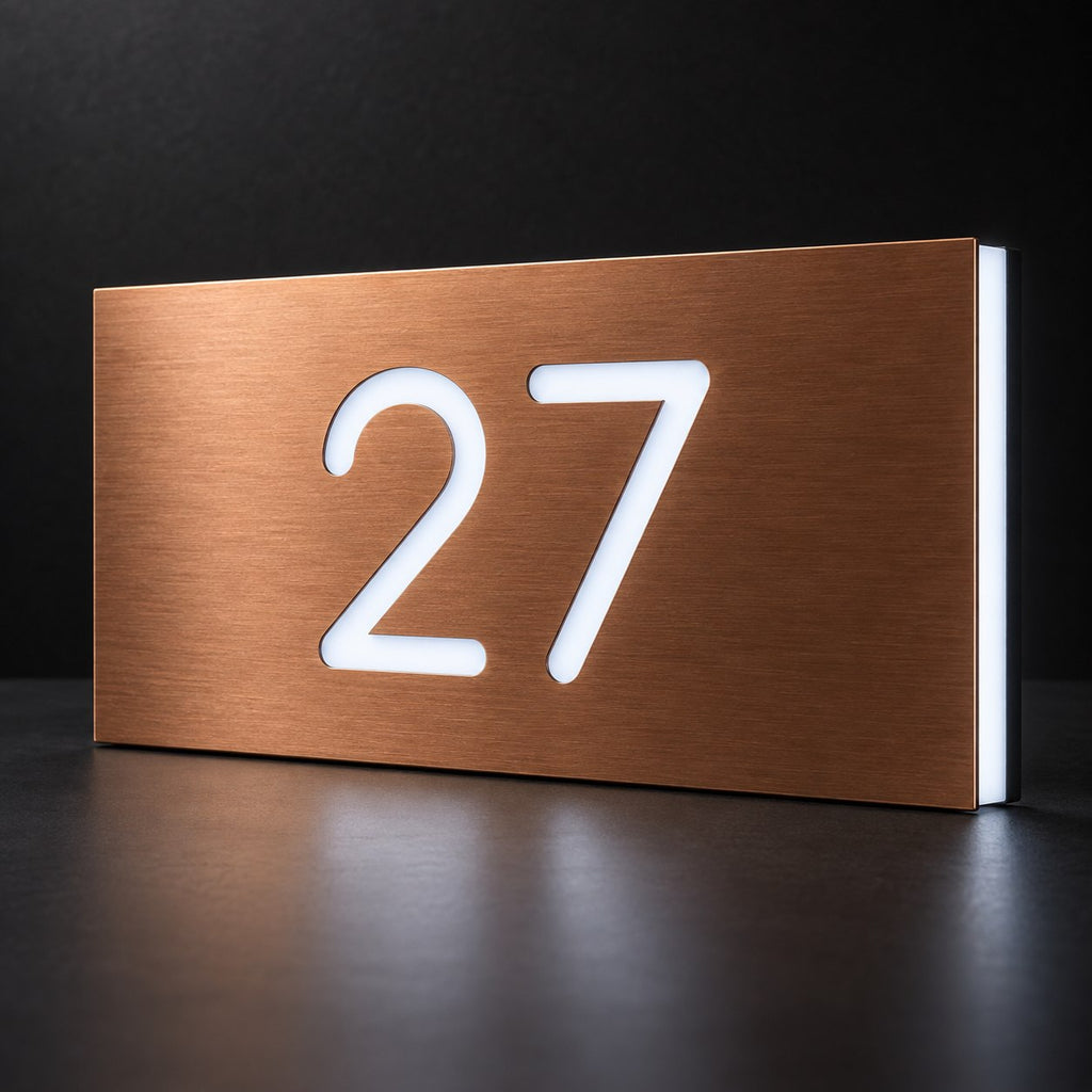 Brushed copper illuminated house number sign with a brushed metal look, The Kawana, displaying 27 shining through in white LED light on a dark reflective surface.