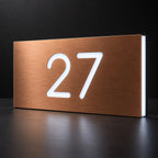 Brushed copper illuminated house number sign with a brushed metal look, The Kawana, displaying 27 shining through in white LED light on a dark reflective surface.