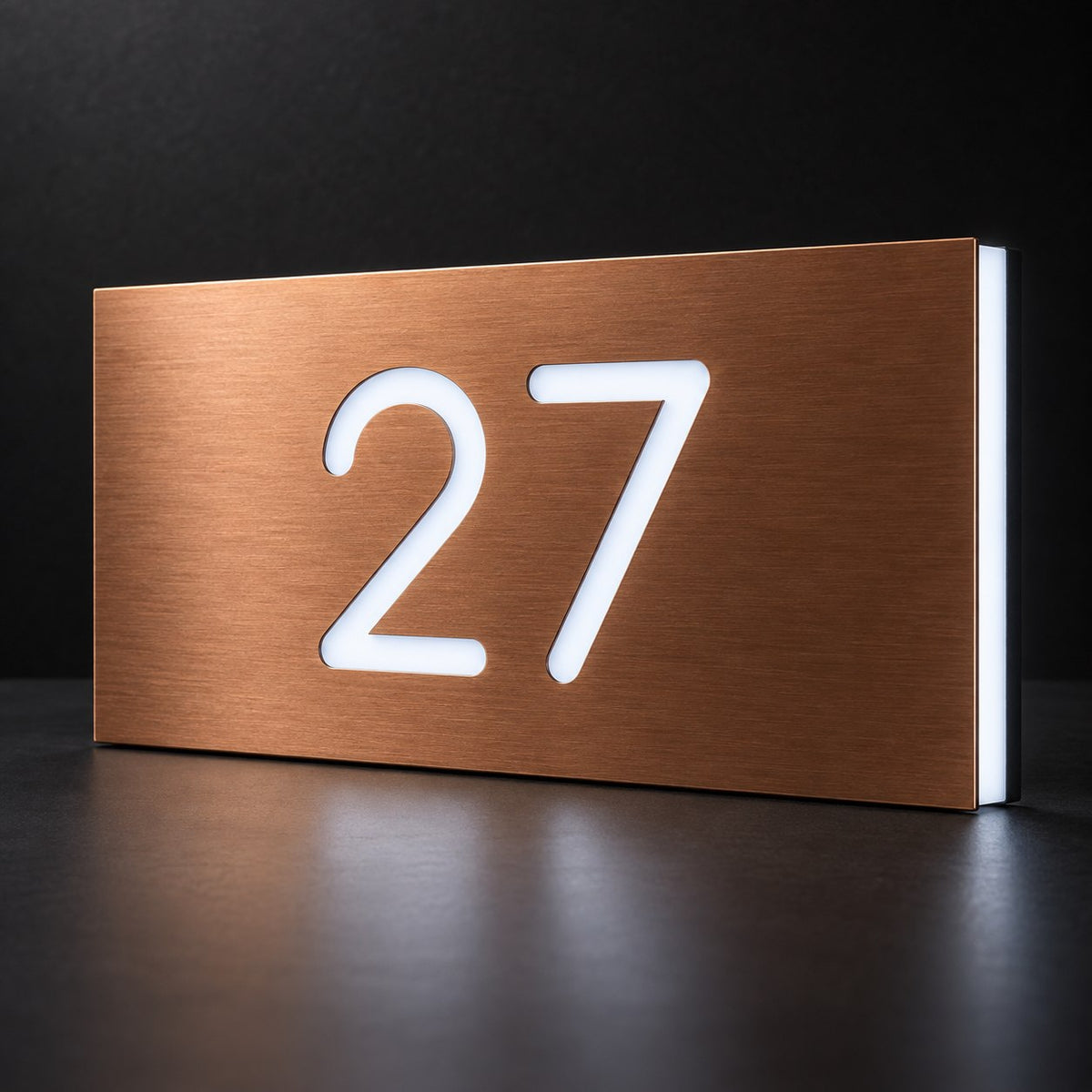 Brushed copper illuminated house number sign with a brushed metal look, The Kawana, displaying 27 shining through in white LED light on a dark reflective surface.