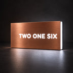 Brushed copper illuminated house number sign with a brushed metal look, The Montville, with the number spelled out as TWO ONE SIX, lit from behind in white.