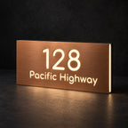 Brushed copper illuminated house number and street name sign with a brushed metal look, The Peregian, 128 Pacific Highway cut through and lit from behind in warm white.