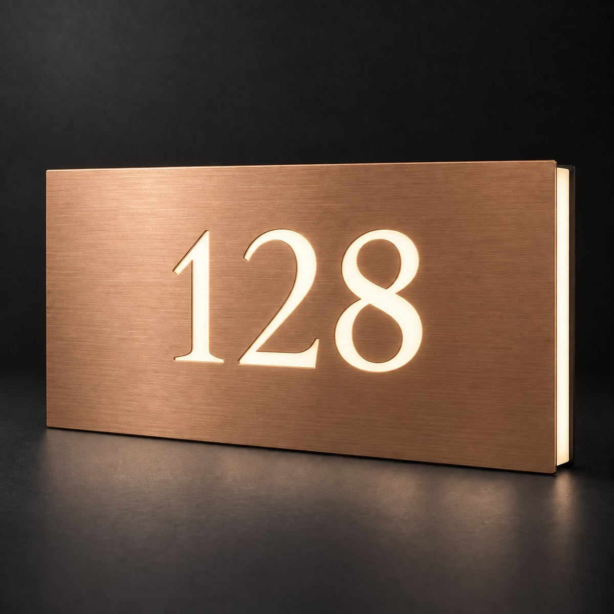 Brushed copper illuminated house number sign with a brushed metal look, The Kawana, displaying 128 in glowing warm white on a dark studio background.