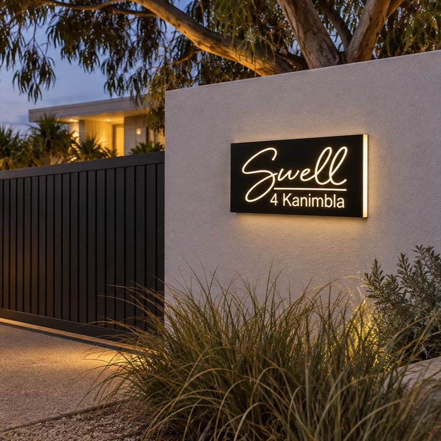 Illuminated house name sign, matte black with warm white script lettering, on a rendered wall beside a slat gate at dusk