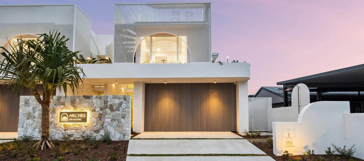 Modern white villa exterior at dusk with a large wooden garage door, stone wall, palm tree, and illuminated sign reading ARCHES