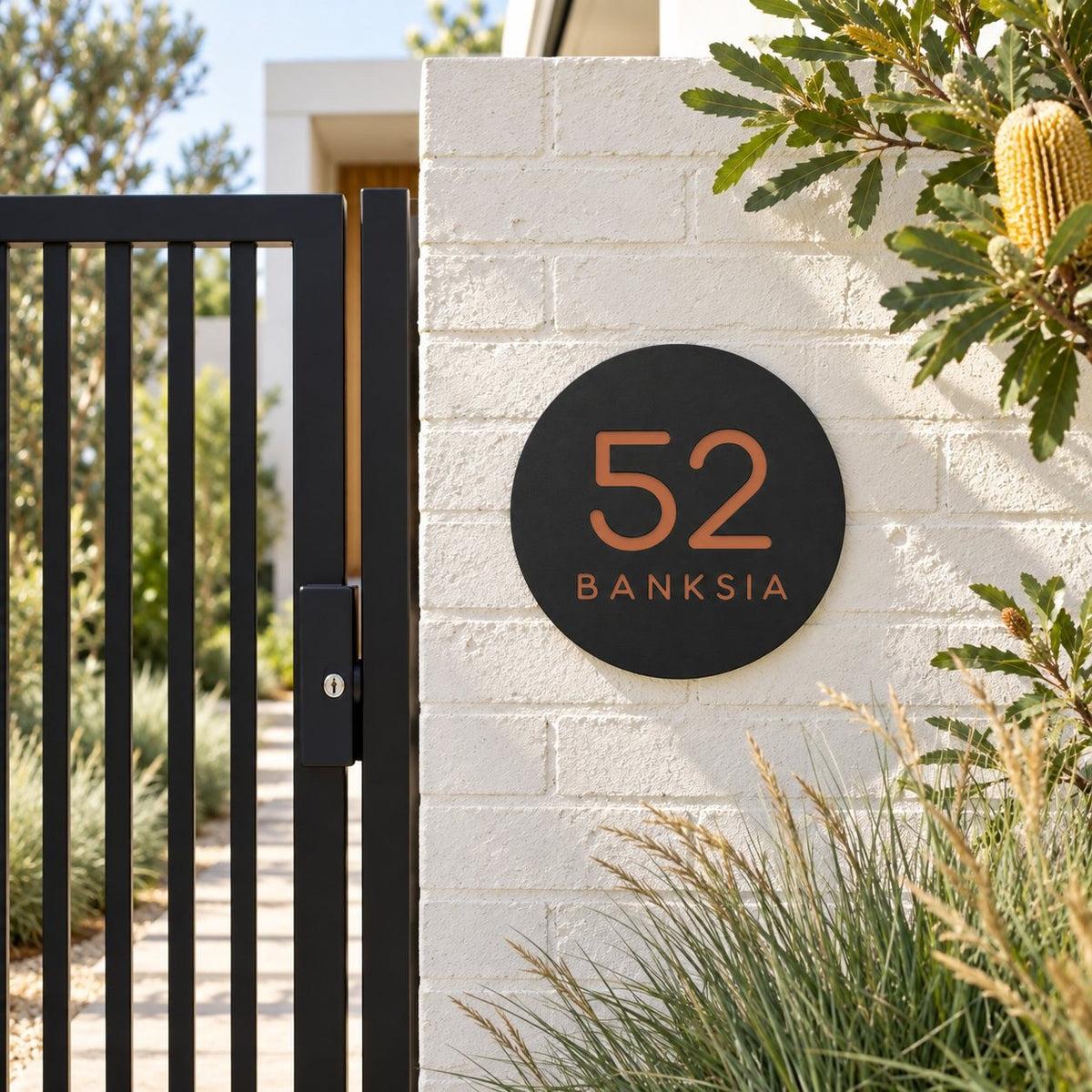 Black round house number and street name sign reading 52 Banksia in brushed copper, The Buderim style, on a white painted brick pillar beside a black gate.