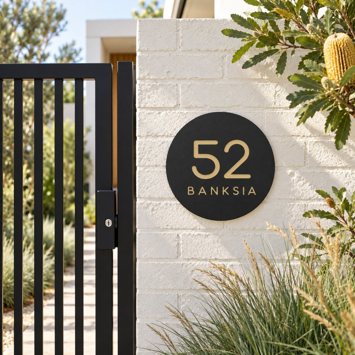Black round house number and street name sign reading 52 Banksia in brushed gold, The Buderim style, on a white painted brick pillar beside a black gate.