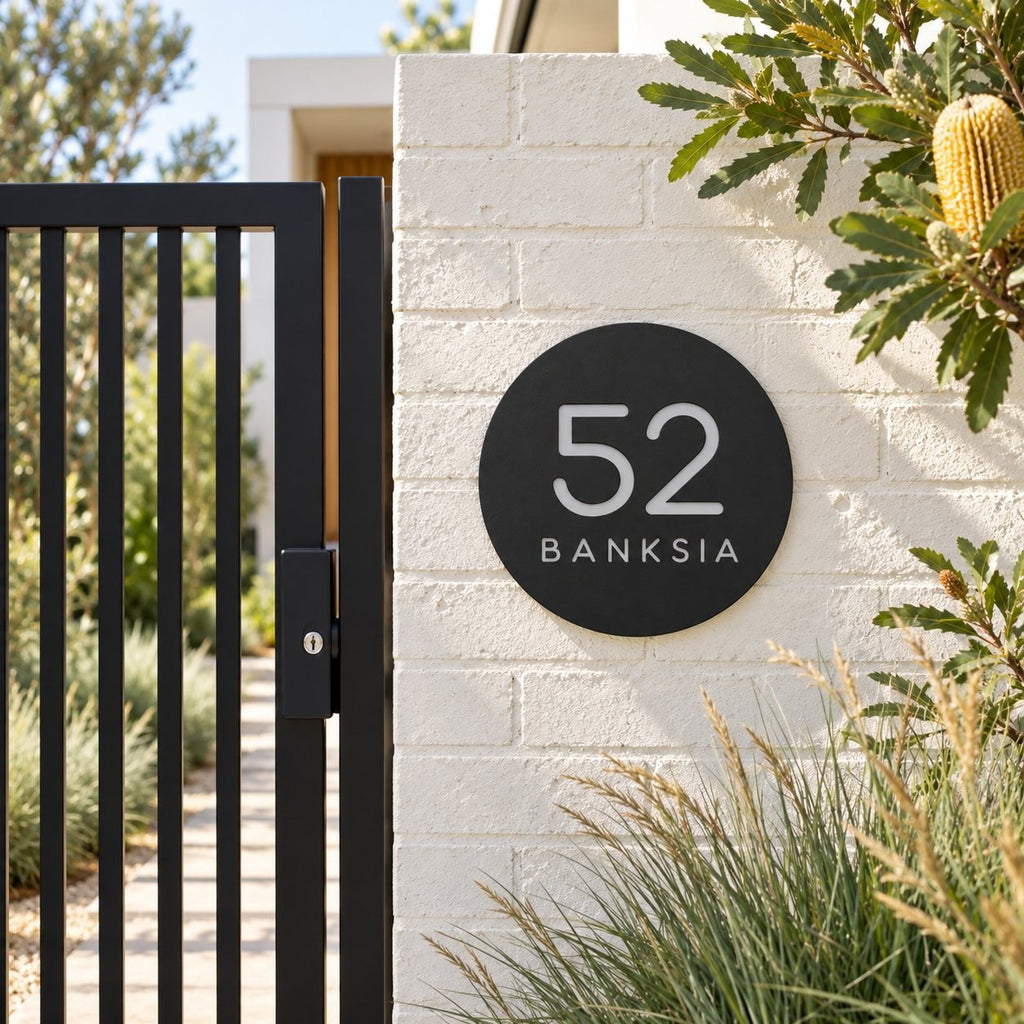 Black round house number and street name sign reading 52 Banksia in brushed silver, The Buderim style, on a white painted brick pillar beside a black gate.