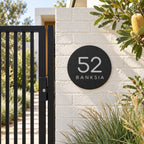 Black round house number and street name sign reading 52 Banksia in brushed silver, The Buderim style, on a white painted brick pillar beside a black gate.