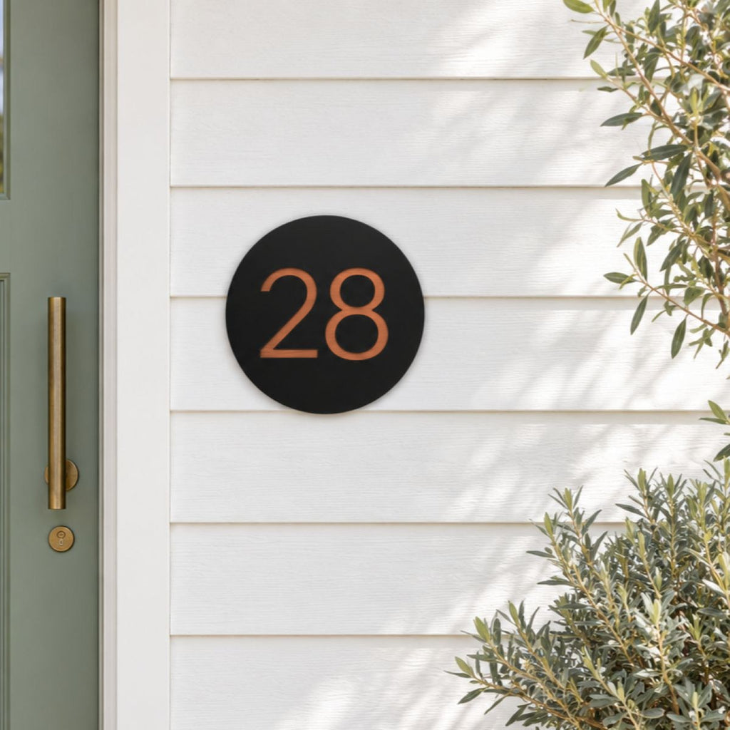 Black round house number sign with the number 28 in brushed copper, The Caloundra style, mounted on white weatherboards beside a sage green door.