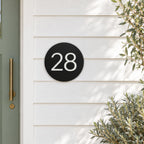 Matte black round house number sign with the number 28 in white, The Caloundra style, mounted on white weatherboards beside a sage green door.