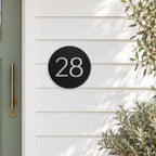 Black round house number sign with the number 28 in brushed silver, The Caloundra style, mounted on white weatherboards beside a sage green door.