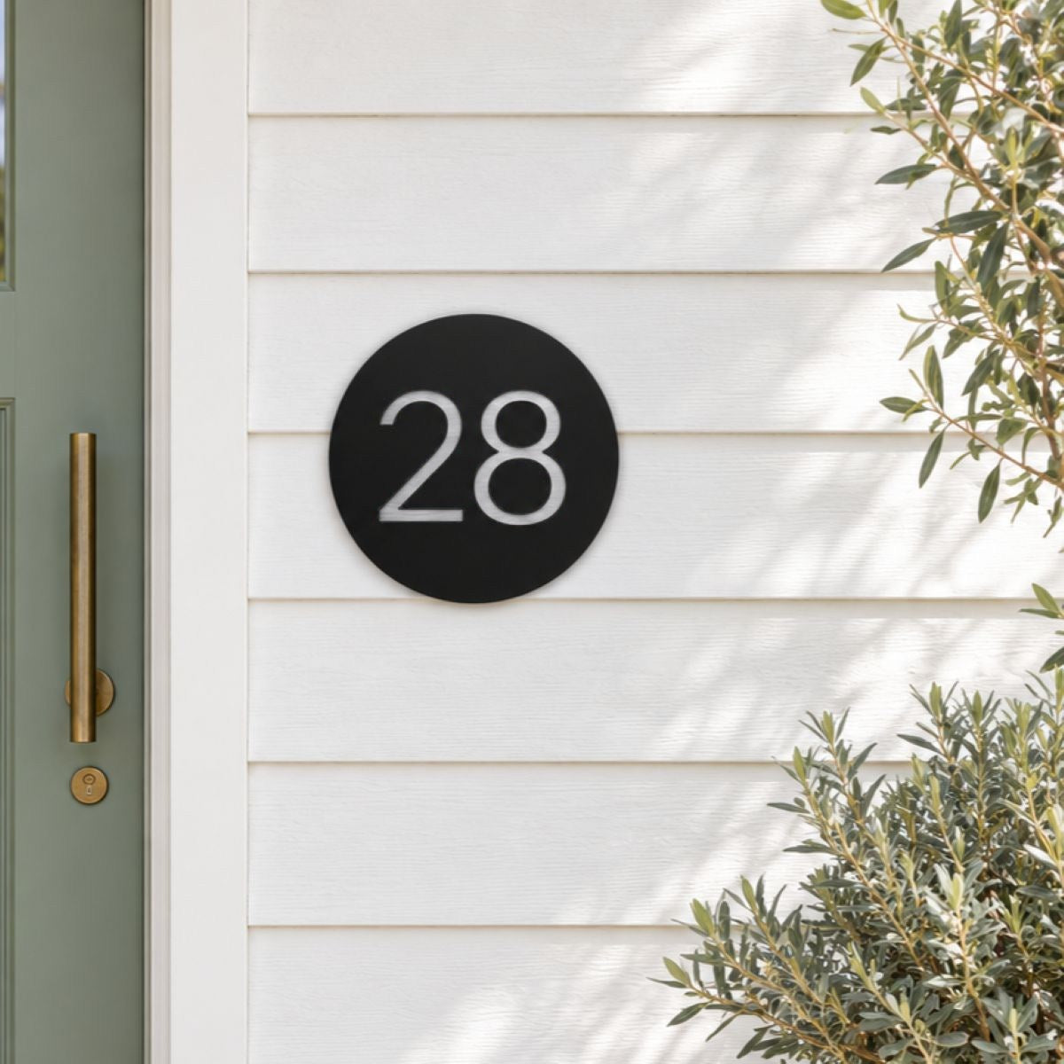 Black round house number sign with the number 28 in brushed silver, The Caloundra style, mounted on white weatherboards beside a sage green door.