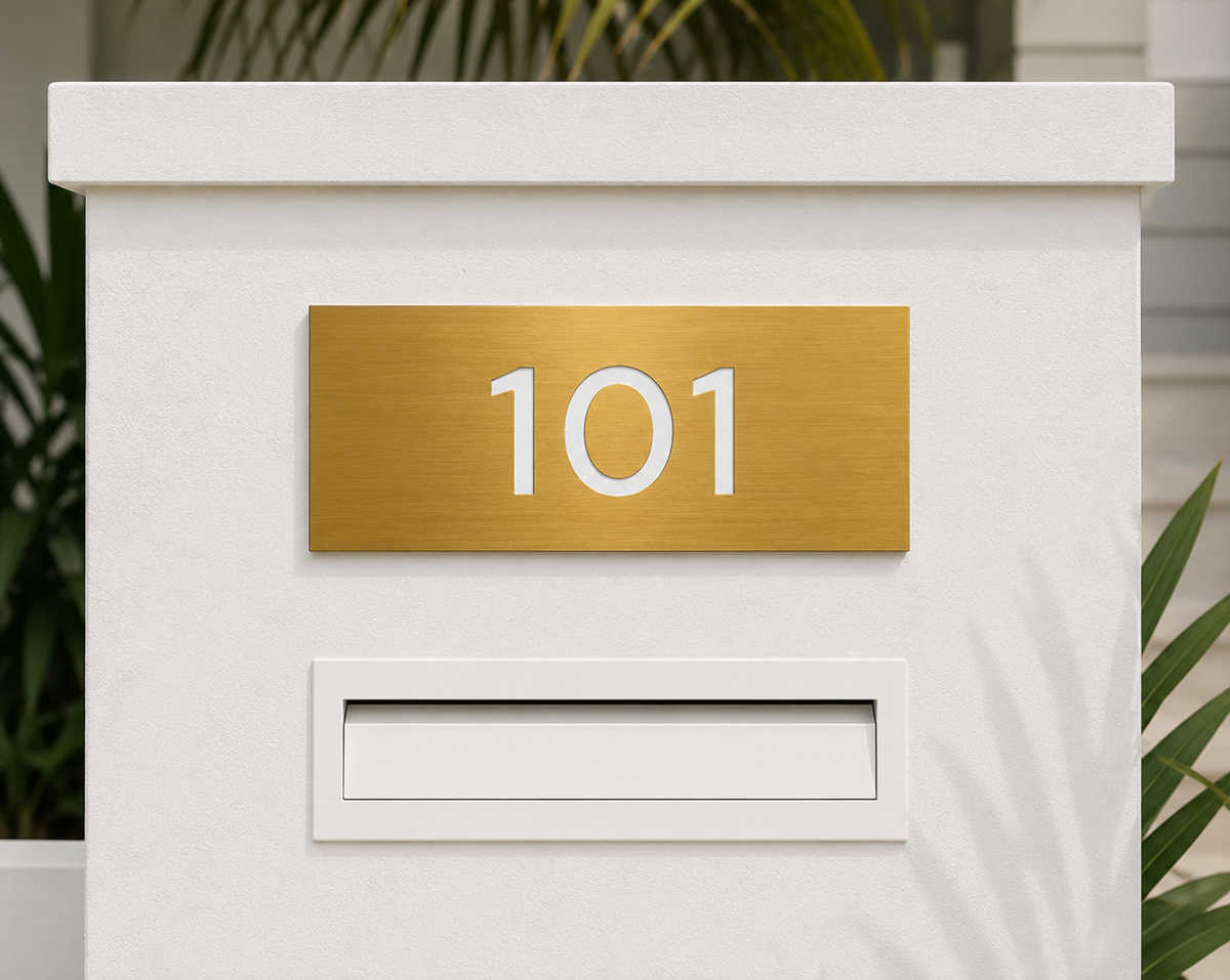 gold metallic house sign with a contrasting white number mounted above a letter box slot on a post box.