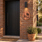 Brushed copper round house number sign with the number 36 and a palm tree cut through to black, The Mooloolaba style, on a red brick wall under a wall light.