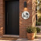 Brushed silver round house number sign with the number 36 and a palm tree cut through to black, The Mooloolaba style, on a red brick wall under a wall light.