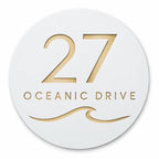 White round house number and street name sign reading 27 Oceanic Drive with a curling wave, cut through to brushed gold, The Mudjimba style, on a plain white background.