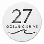 Matte white round house number and street name sign reading 27 Oceanic Drive with a curling wave, cut through to black, The Mudjimba style, on a plain white background.