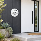 Matte white round house number and street name sign reading 27 Oceanic Drive with a curling wave, cut through to black, The Mudjimba style, on dark vertical cladding beside a white front door.