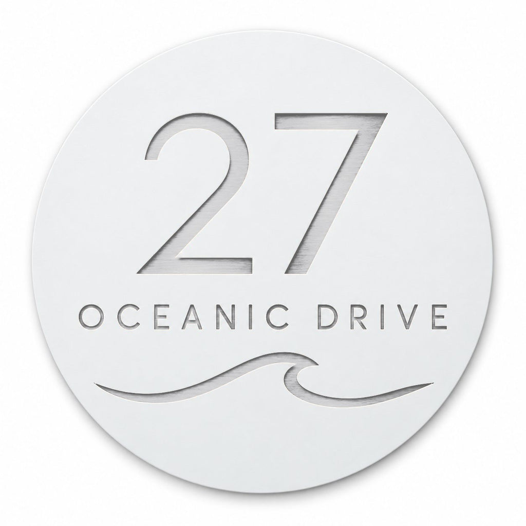 White round house number and street name sign reading 27 Oceanic Drive with a curling wave, cut through to brushed silver, The Mudjimba style, on a plain white background.