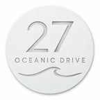 White round house number and street name sign reading 27 Oceanic Drive with a curling wave, cut through to brushed silver, The Mudjimba style, on a plain white background.