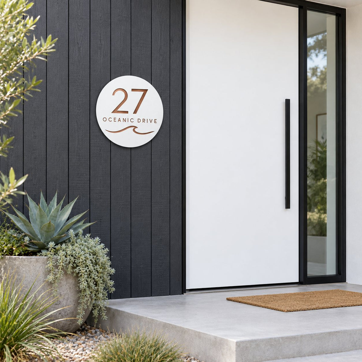 White round house number and street name sign reading 27 Oceanic Drive with a curling wave, cut through to brushed copper, The Mudjimba style, on dark vertical cladding beside a white front door.
