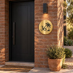 Brushed gold round house number sign with the number 36 and a palm tree cut through to black, The Mooloolaba style, on a red brick wall under a wall light.