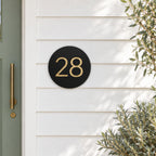 Black round house number sign with the number 28 in gold, The Caloundra style, mounted on white weatherboards beside a sage green door.