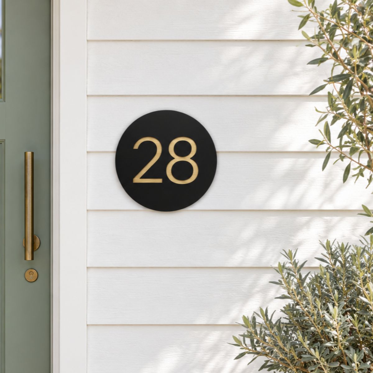 Black round house number sign with the number 28 in gold, The Caloundra style, mounted on white weatherboards beside a sage green door.