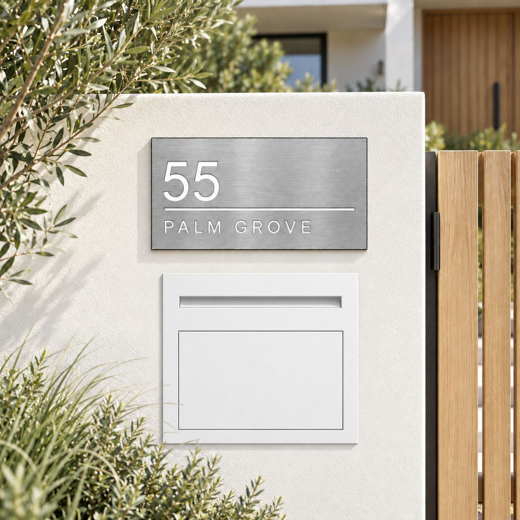 A modern house address plaque and matching mailbox mounted on a stucco wall in front of a wooden gate, with greenery around the property.